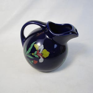 May include: A deep cobalt blue ceramic jug with a rounded, spherical body and a curved handle. The front of the jug features a painted floral design in shades of yellow, white, green, and red, depicting a stylised iris flower.