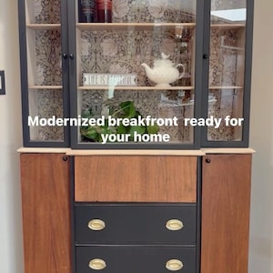 May include: A black and wood breakfront cabinet with glass doors and drawers. The cabinet has a patterned wallpaper backing and gold-colored hardware. The text "LIFE IS BEAUTIFUL" is visible on a small sign inside the cabinet.