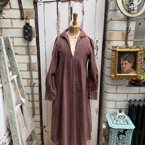 May include: A vintage-style dress in a muted mauve hue, patterned with a small repeating design. The dress has a collared neckline, long sleeves with cuffs, and a flowing shape. It is displayed on a wooden mannequin, showcasing its period style.
