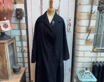 Può includere: Un lungo cappotto nero con colletto e chiusura con bottoni. Il cappotto ha un bordo decorativo lungo i bordi. Il cappotto è esposto su un manichino in un negozio.