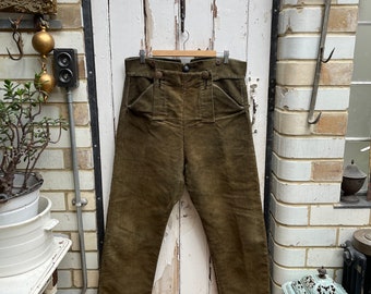 Puede incluir: Un pantalón de pana marrón de cintura alta con varios bolsillos. El pantalón está colgado de una percha de madera contra una puerta de madera blanca.