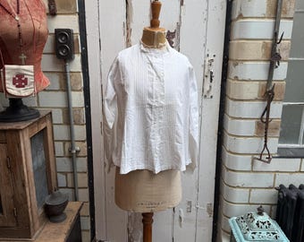 May include: A white cotton blouse with a ruffled collar and a button-up front. The blouse is pleated and has long sleeves. It is displayed on a wooden mannequin against a white brick wall.
