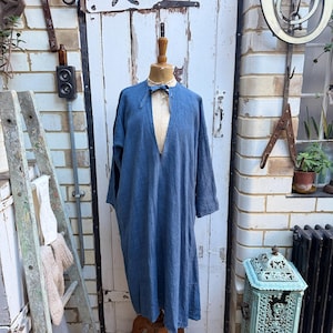 Può includere: Un lungo abito blu con scollo a V profondo è esposto su un manichino di legno. L'abito ha maniche lunghe e una vestibilità ampia. Un piccolo papillon blu scuro è all'altezza dello scollo. Lo sfondo comprende una porta bianca e un muro di mattoni.