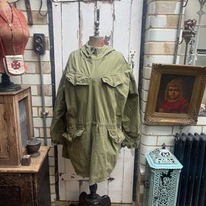Könnte beinhalten: Eine vintage grüne Kapuzenjacke mit Kordelzug in der Taille und mehreren Taschen. Die Jacke befindet sich auf einer Schaufensterpuppe in einem Ladenambiente.