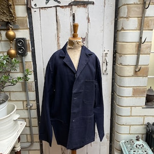 May include: A dark blue work jacket with a button closure and multiple pockets. The jacket is on a wooden mannequin in front of a white painted door.