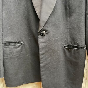 May include: A black tuxedo jacket with a satin lapel and a single button closure. The jacket has two flap pockets on the front.
