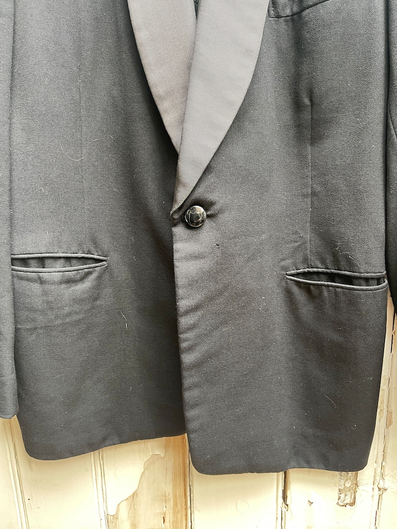 May include: A black tuxedo jacket with a satin lapel and a single button closure. The jacket has two flap pockets on the front.