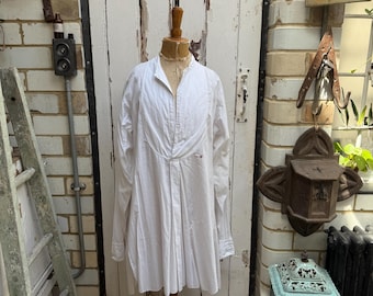 May include: A white long-sleeved shirt with a unique pleated design, displayed on a wooden mannequin. The shirt has a high collar and a button-down front. The mannequin is on a wooden stand. The background includes a white door and brick wall.
