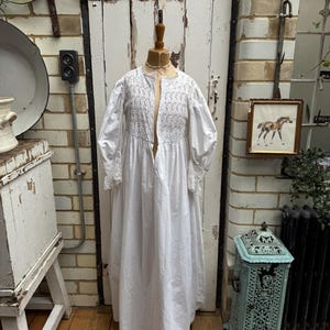 Puede incluir: Camisón largo blanco vintage con detalles de encaje en el corpiño y los puños. El camisón se exhibe en un maniquí contra el telón de fondo de una puerta blanca y una pared de ladrillos. El vestido tiene mangas largas y una falda fluida.
