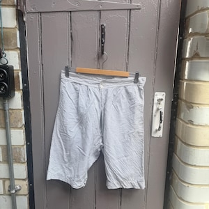 May include: A pair of knee-length shorts featuring a light blue and white pinstripe pattern. The shorts have a button closure at the waistband, a pleated front design, and are suspended from a wooden hanger.