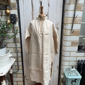 Pode incluir: Uma peça de roupa longa de linho creme com gola alta e fenda lateral, exibida em um manequim de madeira. A peça tem um corte folgado e apresenta um pequeno detalhe bordado em vermelho. O fundo inclui uma porta branca e uma parede de tijolos.