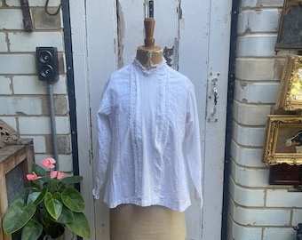 May include: A white cotton blouse with a ruffled collar and long sleeves. The blouse is on a mannequin in front of a white brick wall.