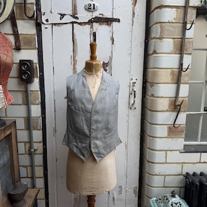 May include: A grey waistcoat with a subtle pattern, displayed on a vintage-style dress form. The form is standing in front of a white wooden door with a black metal number "21" on it.