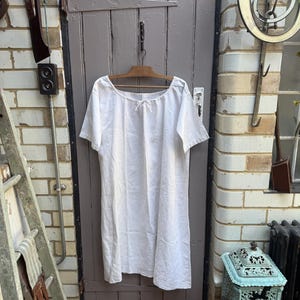 May include: A plain white knee-length shift dress with short sleeves and a wide, rounded neckline featuring a small center tie closure. The garment is constructed from a lightweight fabric and hangs straight from a wooden hanger against a door.