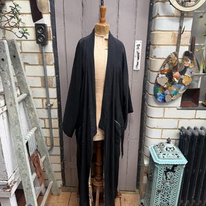 May include: A long, black, open-front robe with wide sleeves, displayed on a beige mannequin. The garment features a solid black exterior and a visible patterned lining along the inner edges, hanging straight down to a mid-calf length.
