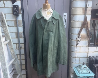 May include: An olive green coat displayed on a wooden mannequin. The coat has a wide collar and a button closure. The mannequin is on a wooden stand. The background includes a gray door, a white brick wall, and a vintage clock.