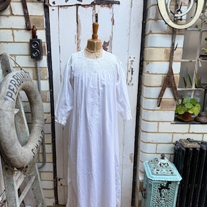 May include: A long, white vintage nightdress with a high neckline and long sleeves. The nightdress is displayed on a mannequin and features delicate embroidery around the neckline and button details down the front. The garment is made of a lightweight fabric.