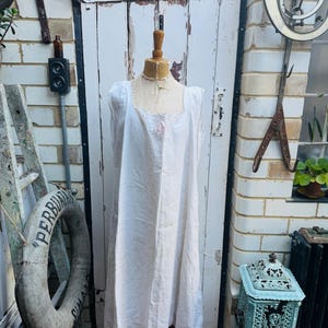 May include: A vintage white linen nightgown displayed on a wooden mannequin. The nightgown features a square neckline with delicate lace trim and short sleeves. The garment is long and flowing, with a simple, elegant design. The background includes a weathered white door and brick wall.
