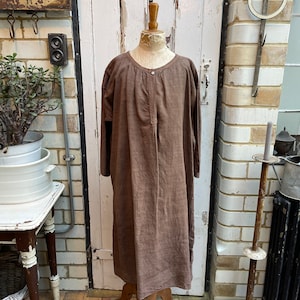 May include: A brown linen dress with long sleeves and a button-up front. The dress is displayed on a mannequin against a white brick wall.