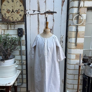 May include: A white linen nightgown with a lace trim around the neckline and sleeves. The nightgown is displayed on a mannequin against a white brick wall.