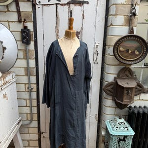 Può includere: Un lungo vestito blu scuro con scollo a V è esposto su un manichino di legno. L'abito ha maniche lunghe e una vestibilità ampia. Lo sfondo include una porta bianca, un muro di mattoni e oggetti decorativi.