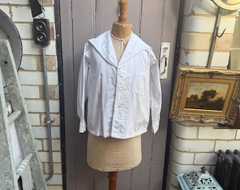 May include: A white long-sleeved shirt with a wide sailor-style collar, a front button closure, and a single chest pocket. The shirt is displayed on a beige fabric dress form with a wooden stand, featuring a visible neck opening and cuffs.