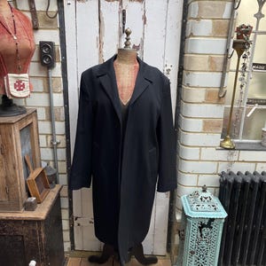 May include: A long, black coat on a vintage dressmaker's dummy. The coat features a classic lapel and a tailored cut. The dummy is positioned in front of a white door and brick wall, creating a vintage aesthetic.