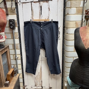 May include: A pair of black cotton pants hanging on a wooden hanger. The pants have a button fly and a wide waistband. The pants are hanging in front of a white wooden door with a number "21" on it.
