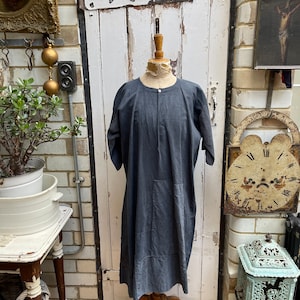 May include: A gray linen dress with a round neckline and a button closure. The dress has a relaxed fit and a slightly A-line silhouette. It is displayed on a mannequin against a white brick wall.