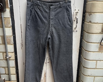 Puede incluir: Pantalón gris a rayas con cierre de botón y dos bolsillos delanteros. El pantalón está colgado de una percha de madera contra una puerta de madera blanca.