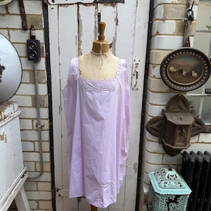 Può includere: Una camicia da notte vintage, lilla chiaro, su un manichino di legno. La camicia da notte ha uno scollo quadrato con dettagli ricamati. Lo sfondo comprende una porta bianca, un muro di mattoni e oggetti decorativi.