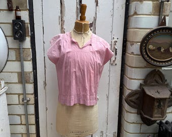 May include: A light pink, short-sleeved blouse displayed on a vintage dress form. The blouse has a V-neck and a textured, vertical striped pattern. The dress form is beige and mounted on a wooden stand. The background includes a white door and brick wall.