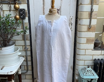 May include: A white sleeveless linen dress displayed on a wooden mannequin. The dress features a simple design with a round neckline and a button fastening at the neck. The mannequin is positioned on a wooden stand against a white brick wall.