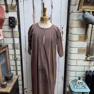 May include: A brown, long-sleeved garment with a button closure at the neck. The garment is displayed on a mannequin against a white brick wall.