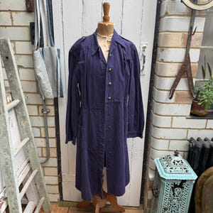 May include: A long, dark blue dress coat with gold buttons is displayed on a wooden mannequin. The coat has long sleeves and a button-down front. The coat is made of a soft, flowing fabric. The mannequin is on a wooden stand.