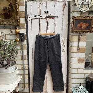 Può includere: Pantaloni da lavoro grigi con toppe appesi a una porta di legno con il numero "21". I pantaloni hanno un aspetto usurato con toppe e cuciture visibili.