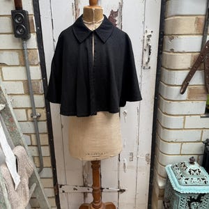 May include: A black cape with a collar is displayed on a vintage dress form. The cape has a textured appearance and a simple, classic design. The dress form is made of tan fabric and is mounted on a wooden stand.