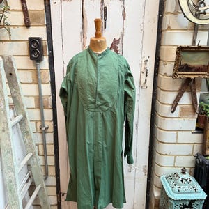 May include: An olive green garment with a mandarin collar and buttoned placket, displayed on a wooden dress form. The garment has long sleeves and a pleated front. The background includes a white door and a vintage aesthetic.