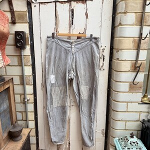 May include: A pair of gray cotton pants with white patches on the knees. The pants are hanging on a wooden hanger against a white wooden door.