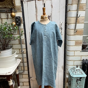 Op de afbeelding: Een lichtblauwe linnen jurk met een knoopsluiting en korte mouwen. De jurk wordt getoond op een mannequin tegen een witte bakstenen muur.