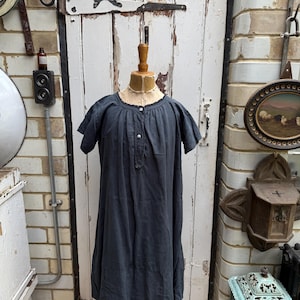 May include: A dark gray, short-sleeved dress on a wooden mannequin. The dress has a round neckline, button details, and a loose, flowing silhouette. The background includes a white door with the number 21, brick walls, and vintage decor.