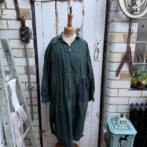 May include: A dark green dress with long sleeves and a collared neckline, displayed on a wooden mannequin. The dress features a dark blue patch. The background includes a white door and a brick wall.