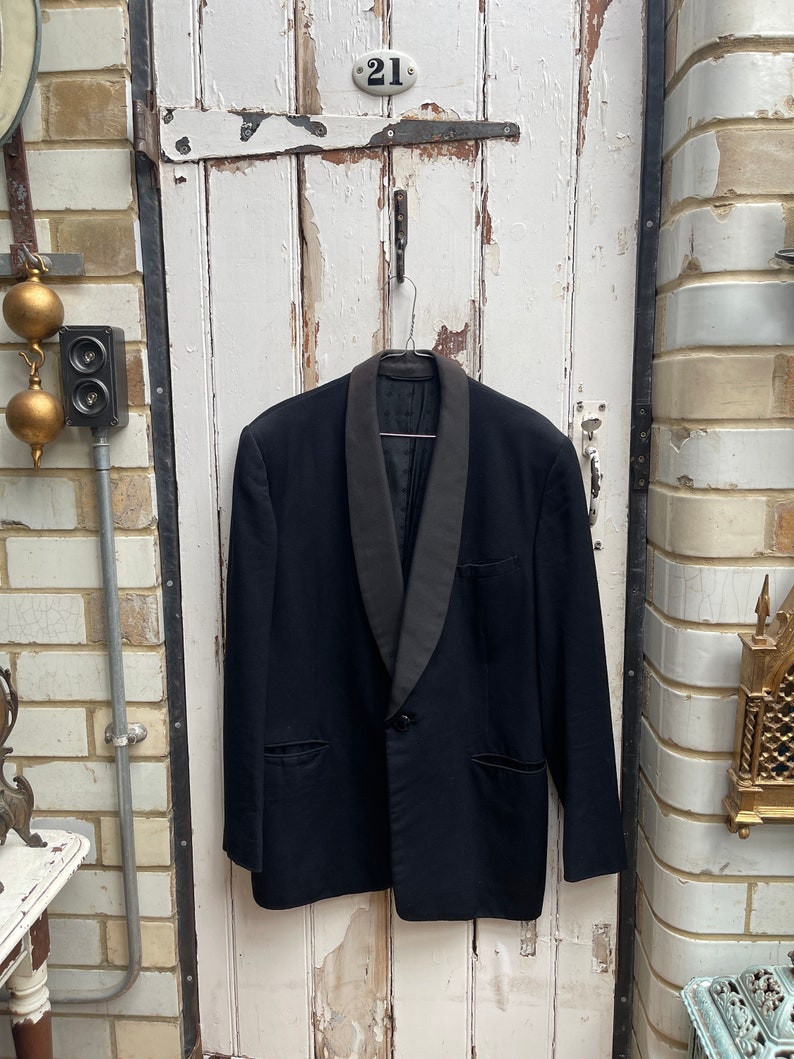 May include: A black tuxedo jacket with a satin shawl collar. The jacket is hanging on a hook against a white wooden door.