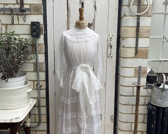 Op de afbeelding: Een witte vintage jurk met een gerimpelde halslijn en een gelaagde rok. De jurk is op een mannequin voor een witte bakstenen muur.