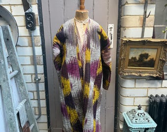 May include: A long-sleeved, quilted robe featuring a vibrant ikat pattern in shades of mustard yellow, deep purple, and grey. The garment has an open front, a V-neckline, and a straight silhouette, displayed on a wooden mannequin torso.