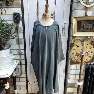 May include: A long, loose-fitting, grey linen dress with a round neckline and a drawstring tie at the neck. The dress is displayed on a mannequin against a white brick wall.