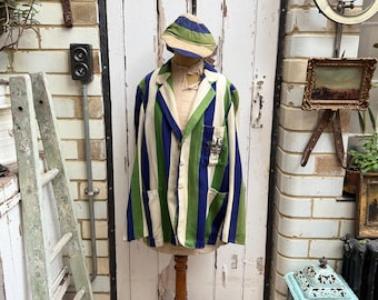 Può includere: Un blazer vintage a righe blu, verde e crema, esposto su un manichino di legno. Il blazer ha un design classico con taschino e berretto a righe abbinato. Lo sfondo comprende una porta bianca e decorazioni antiche.