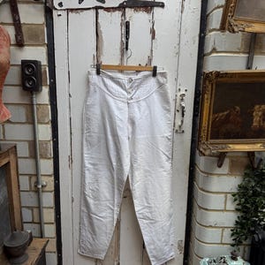 Könnte beinhalten: Weiße Hose mit hohem Bund, die an einem Holzbügel hängt. Die Hose hat einen Knopf- und Reißverschluss, mit sichtbaren Nahtdetails. Der Hintergrund ist eine verwitterte weiße Tür.