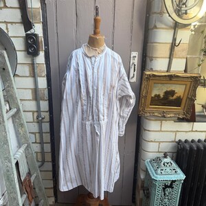 May include: A long-sleeved, knee-length nightshirt featuring a white base with vertical tan and brown stripes. The garment has a button-down placket at the center chest, a small mandarin collar, and a loose, flowing fit displayed on a wooden mannequin.