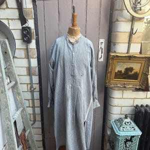 May include: A long-sleeved, light blue shirt with a mandarin collar and a partial button-down front. The garment features a loose, flowing fit, long sleeves with cuffs, and a visible diagonal seam detail on the lower front section of the fabric.